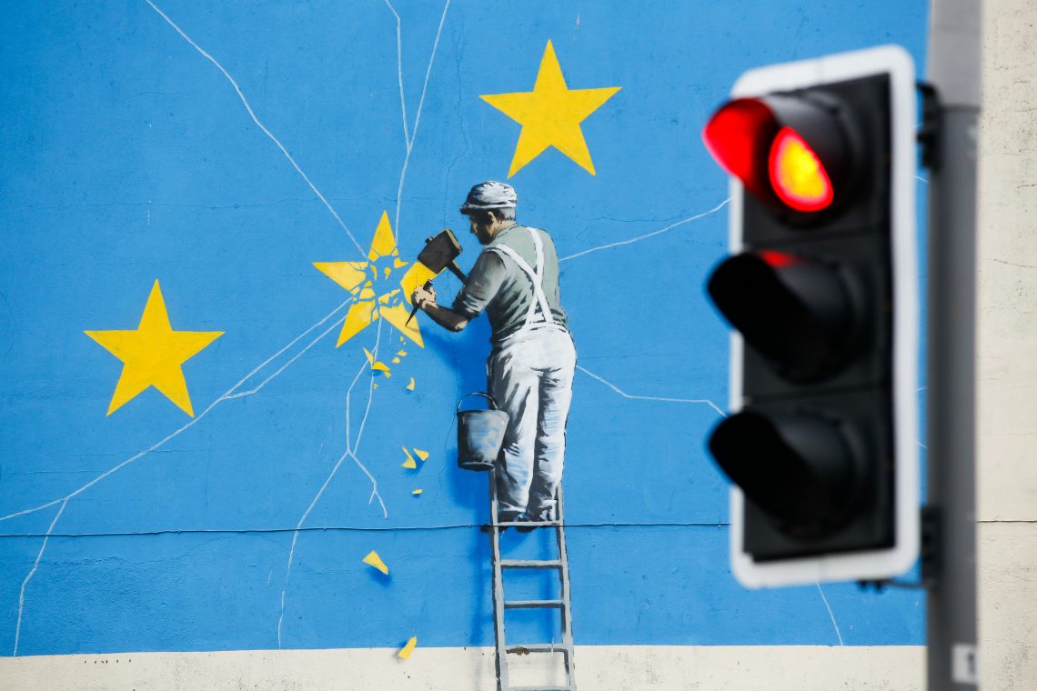 Vor einem Banksy Wandbild, auf dem ein Arbeiter einen EU-Stern aus der Flagge meißelt, leuchtet eine rote Ampel