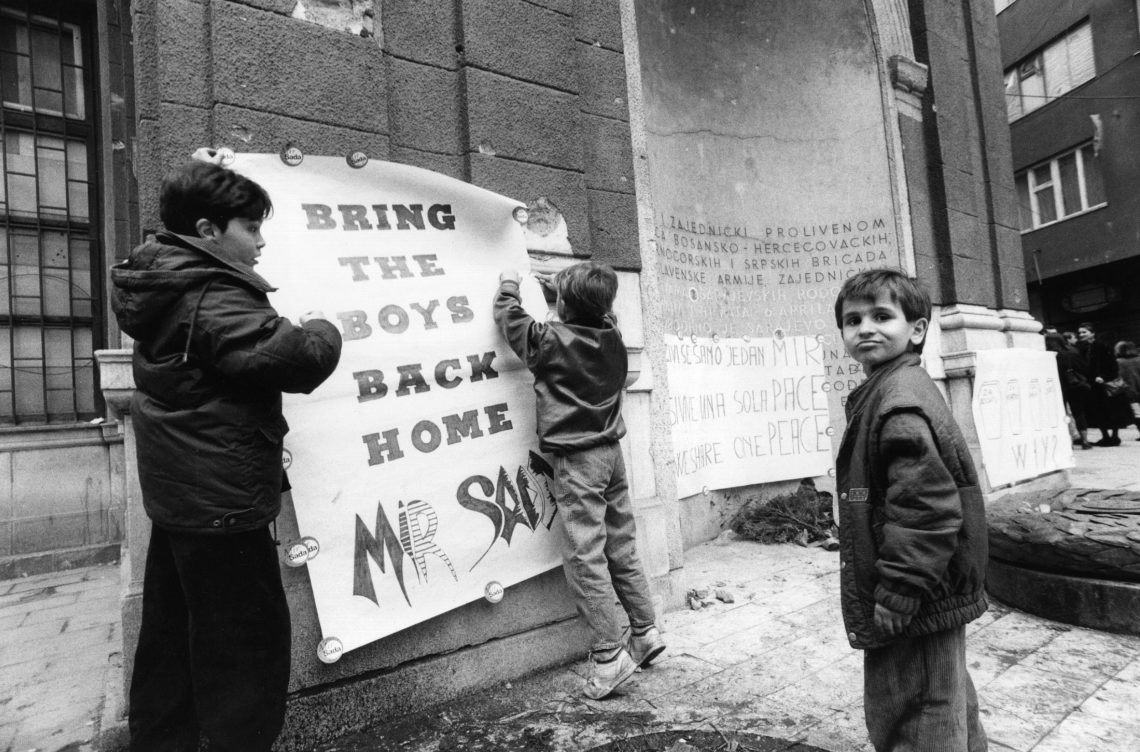 Jungen im Bosnienkrieg, die ein Poster mit der Aufschrift Bring the boys back home an einer Hauswand anbringen