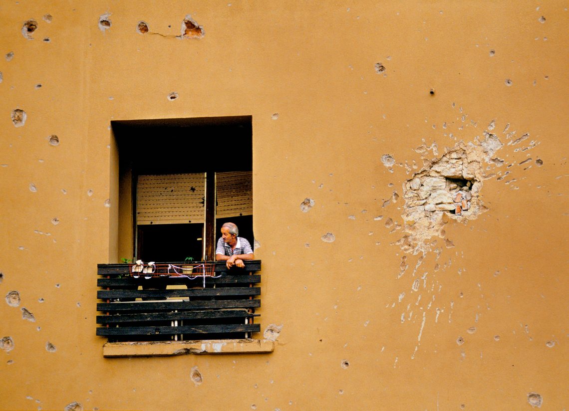 Mann schaut aus Wohnung in Mostar, die mit Einschlaglöchern übersät ist