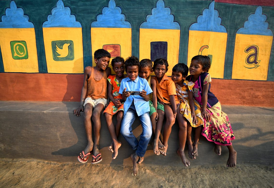 Indische Kinder schauen einen Cartoon auf einem Handy vor einer Wand, die mit Social Media Symbolen bemalt ist