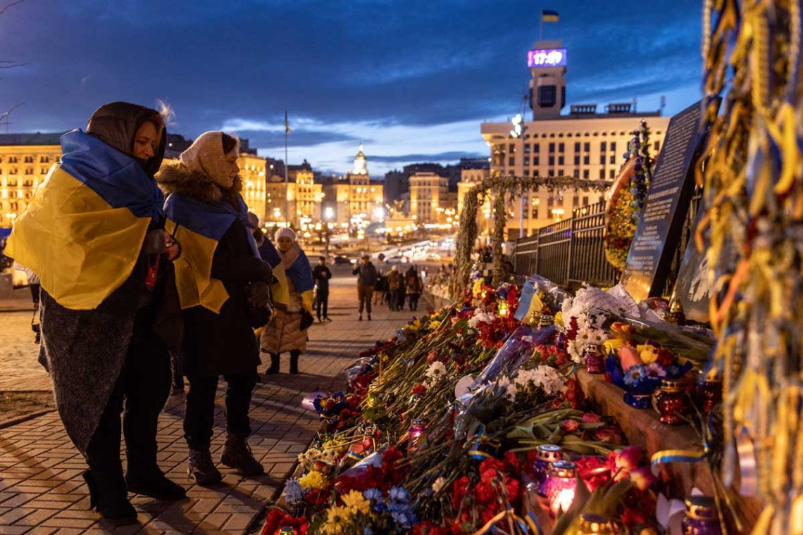 Frauen, gehüllt in ukrainische Flaggen, blicken auf Blumen und Kränze, die im Gedenken an die Toten des Euromaidan niedergelegt wurden. Kiev, Ukraine, im Februar 2022.