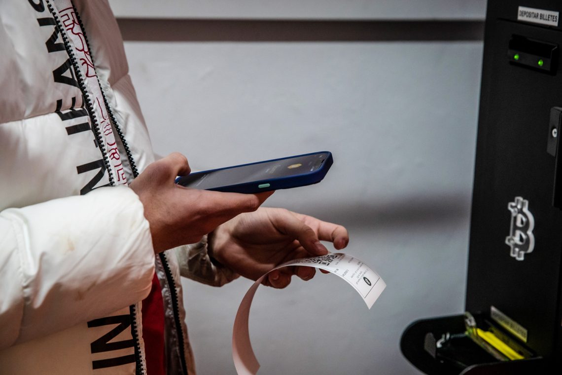 Folgen einer Cyberattacke: Foto einer Frau die mit einem Handy einen Beleg an einem Bitcoin-Bankomaten überprüft.