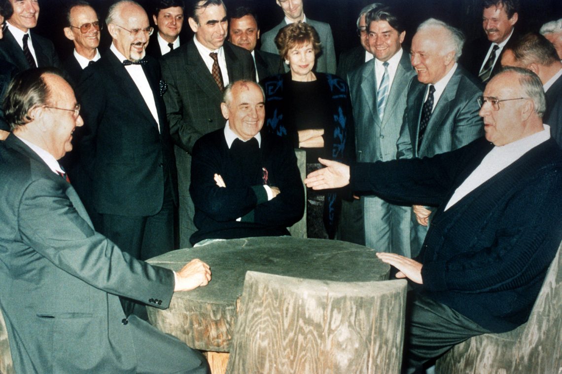 Foto aus dem Jahr 1990: Der deutsche Außenminister Hans-Dietrich Genscher, der Präsident der Sowjetunion, Mikhail Gorbatschow und der deutsche Bundeskanzler Helmut Kohl sitzen gemeinsam an einem rustikalen Tisch auf der Regierungsdatscha Gorbatschows.