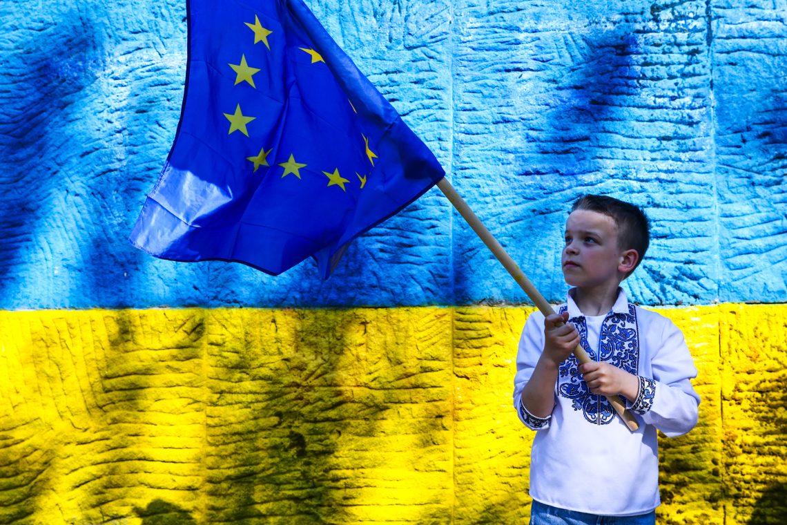 Die Ukraine und die EU: Foto eines Jungen, der vor einer Wand in den Farben der Ukraine eine Fahne der EU schwenkt.