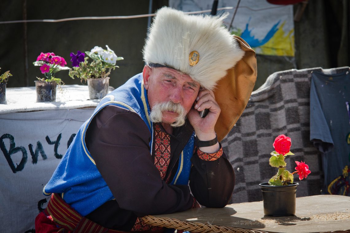 Russland und die Ukraine: Foto eines Mannes mit Kosakenmütze und einer Weste in den Nationalfarben der Ukraine sitzt telefonierend an einem Tisch.