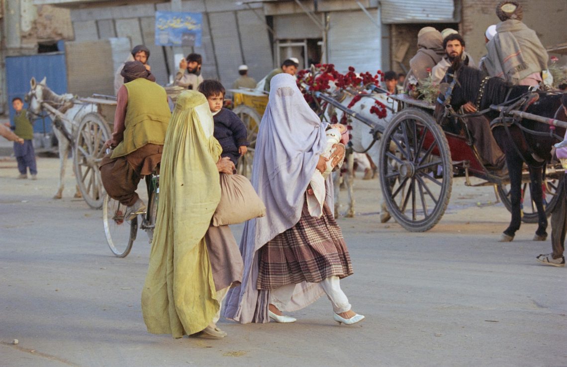 Foto einer Straßenszene in Afghanistan, Im Vordergrund Frauen mit Burkas über moderner Kleidung mit Kindern auf dem Arm, im HIntergrund von Pferden gezogene Wagen.