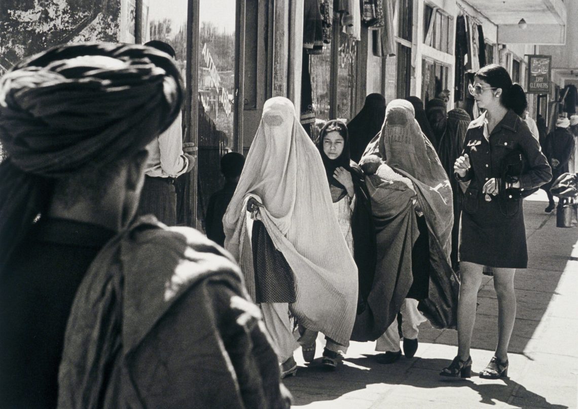 Foto einer Straßenszene in Kabul in den 1970er Jahren: Frauen in Burkas und Minikleid, im Vordergrund ein Mann mit traditionellem Tuch.