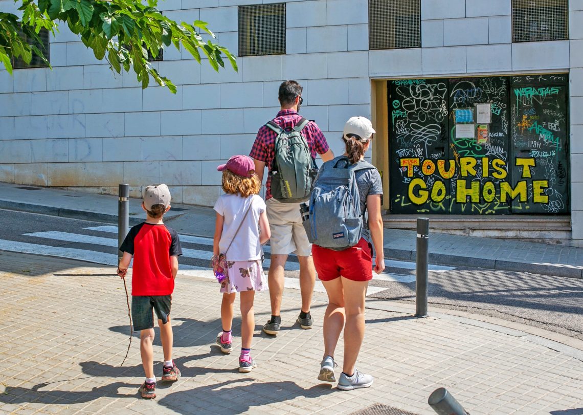 Touristen in Massen: Foto einer Straßenszene mit einer Familie im Urlaub, die an einem Haus ein Grafitti "Tourist go Home" liest.