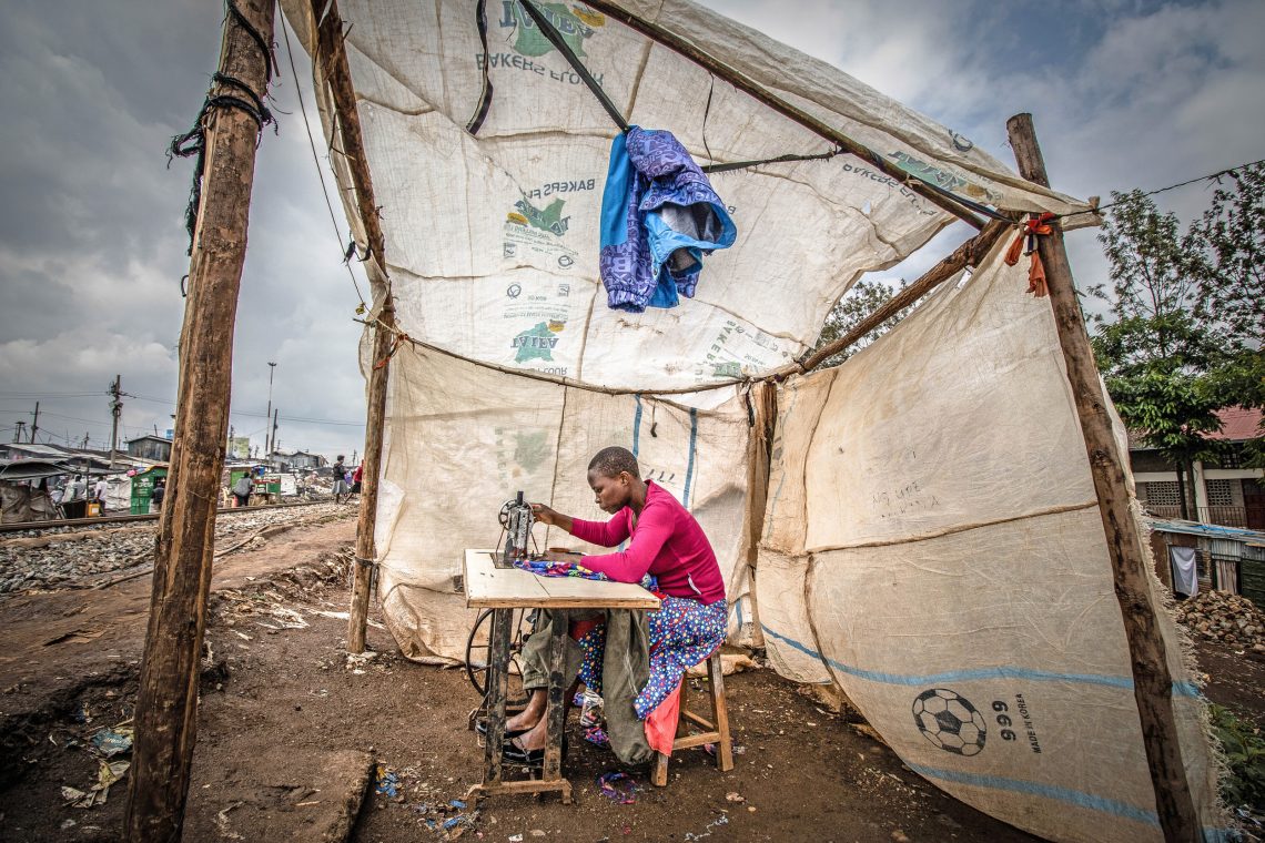 Afrika Bevölkrungswachstum: Eine junge Frau sitzt an einer Nähmaschine unter Stoffplanen an einer unbefestigten Straße. Schneiderin im Kibera-Slum in Nairobi, Kenia, 2021
