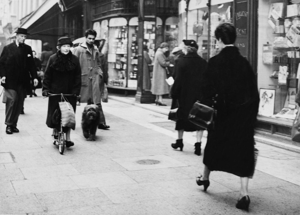 Schwarzweiß Foto von einer belebten Straße mit vielen Fußgängern und großen Schaufenstern. Eine ältere Frau ist mit einem Roller unterwegs, an dem eine Einkaufstasche hängt. Neben ihr läuft ein mittelgroßer Hund mit längeren Haaren.