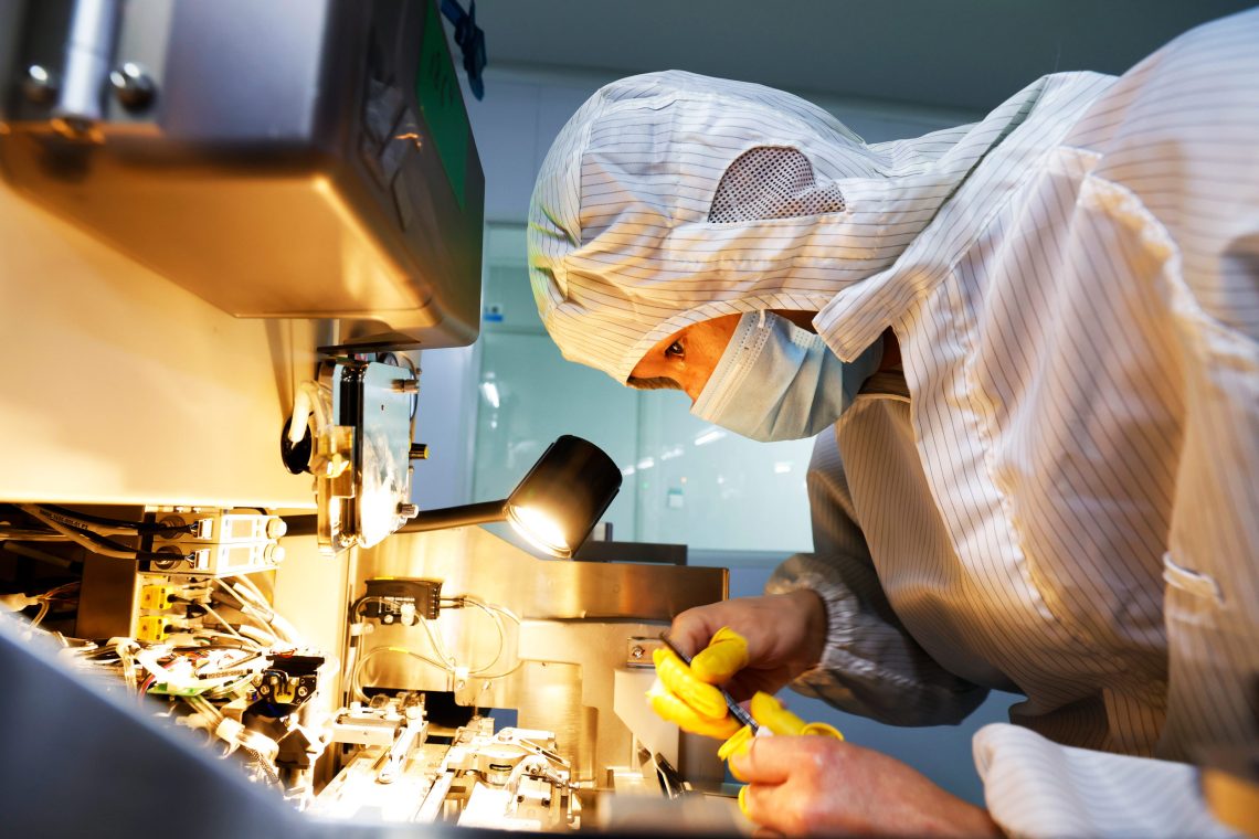 Eine junge Frau in weißer Schutzkleidung beugt sich über einen Arbeitsplatz elektronischen Schaltkreisen und Mikroskopen.