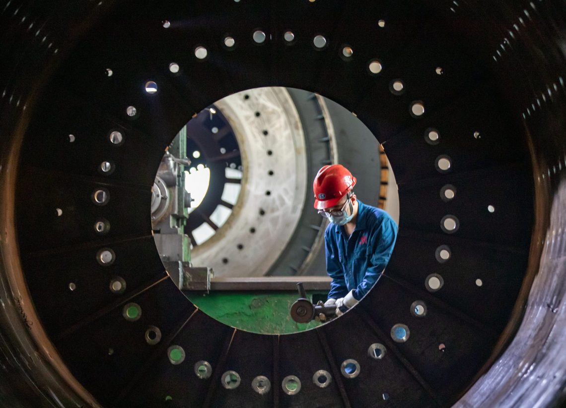 Foto einer kreisförmigen Stahlkonstruktion in China, die von einem Arbeiter im Blaumann und mit rotem Helm mit einer Flex bearbeitet wird.