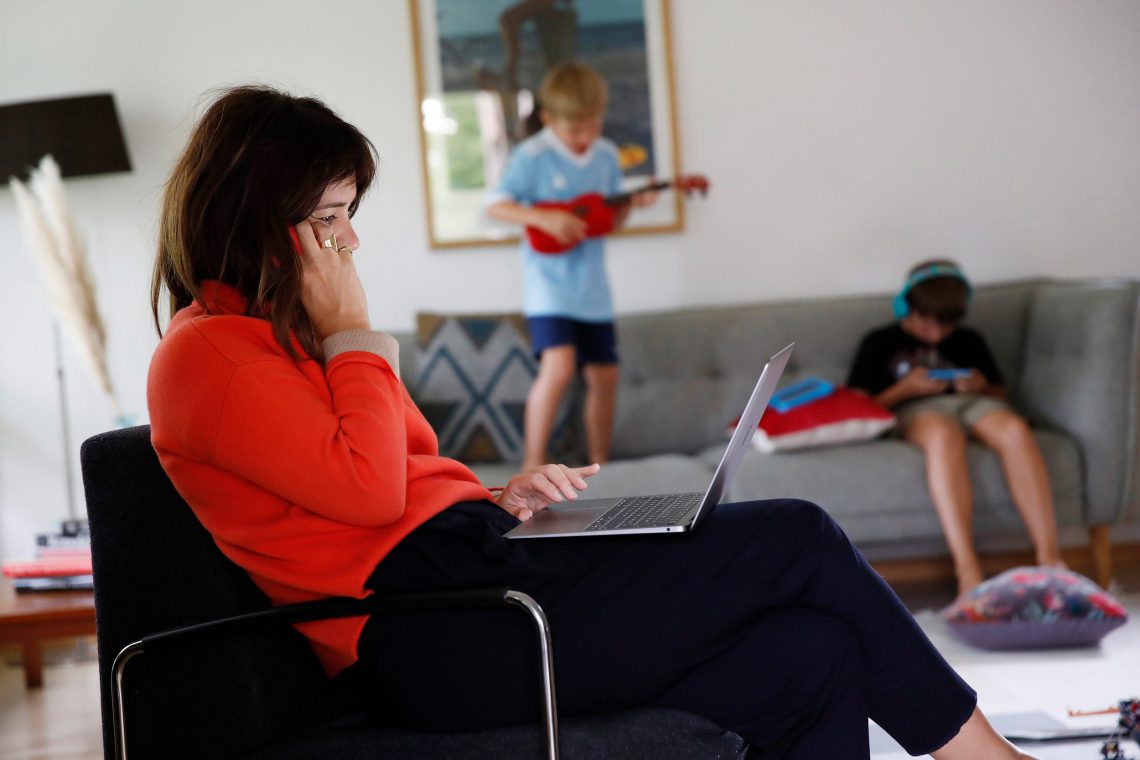 Eine Frau sitzt in einem Wohnzimmer in einem Sessel udn telefoniert mit einem Mobiltelefon. Auf ihren Knien ist ein aufgeklappter Laptop. Im Hintergrund spielen Kinder.