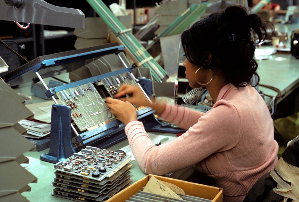 Eine Frau mit einem rosafarbenen Pullover sitzt an einem Arbeitsplatz. Neben ihr liegen Festplatten für einen Computer und sie montiert elektronische Teile auf ein Mainboard.