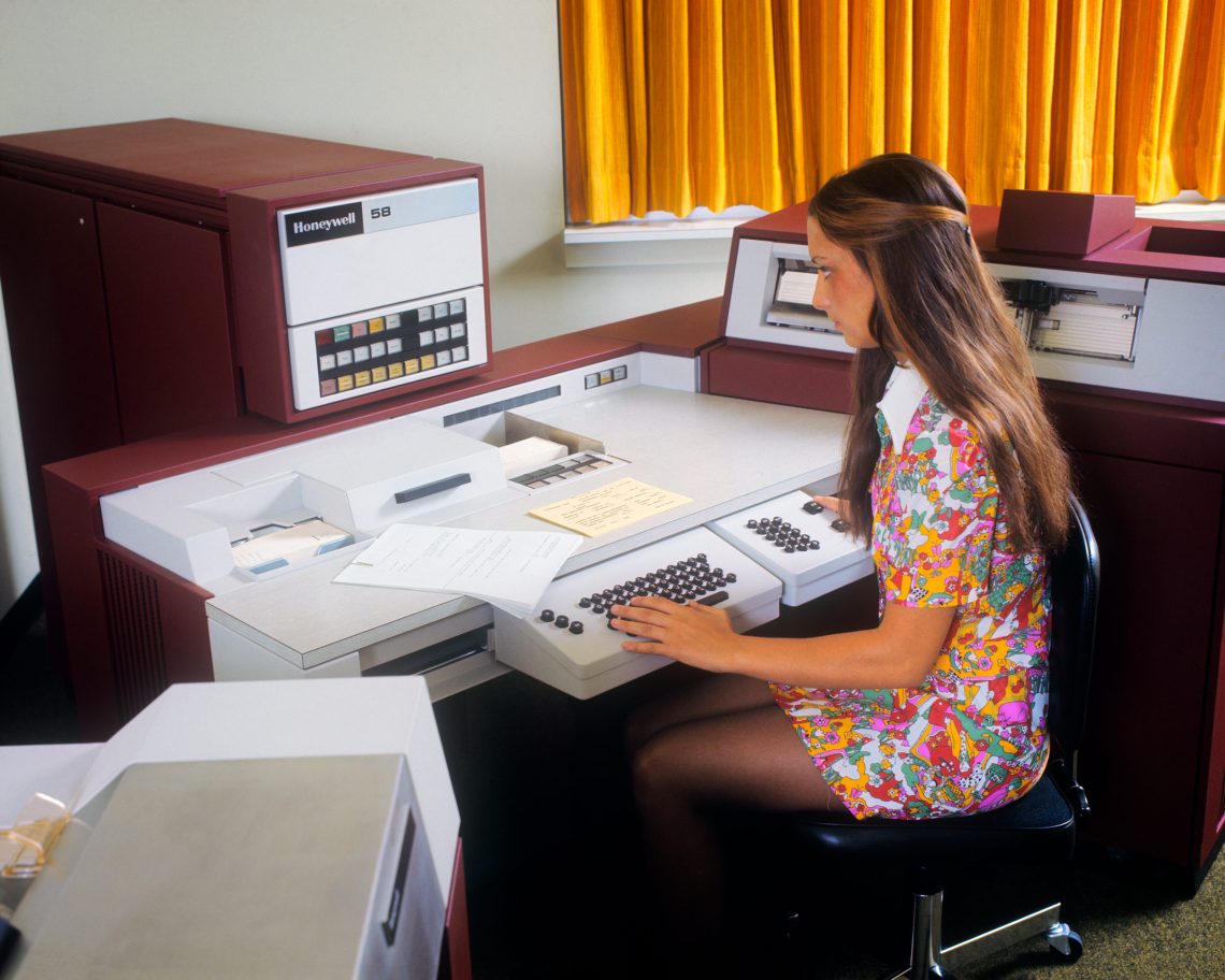 Arbeit: Eine Frau mit langen Haaren und einem gebluemten Kleid sitzt an einer großenn Rechenmaschine an einem Schreibtisch mit Tastatur.