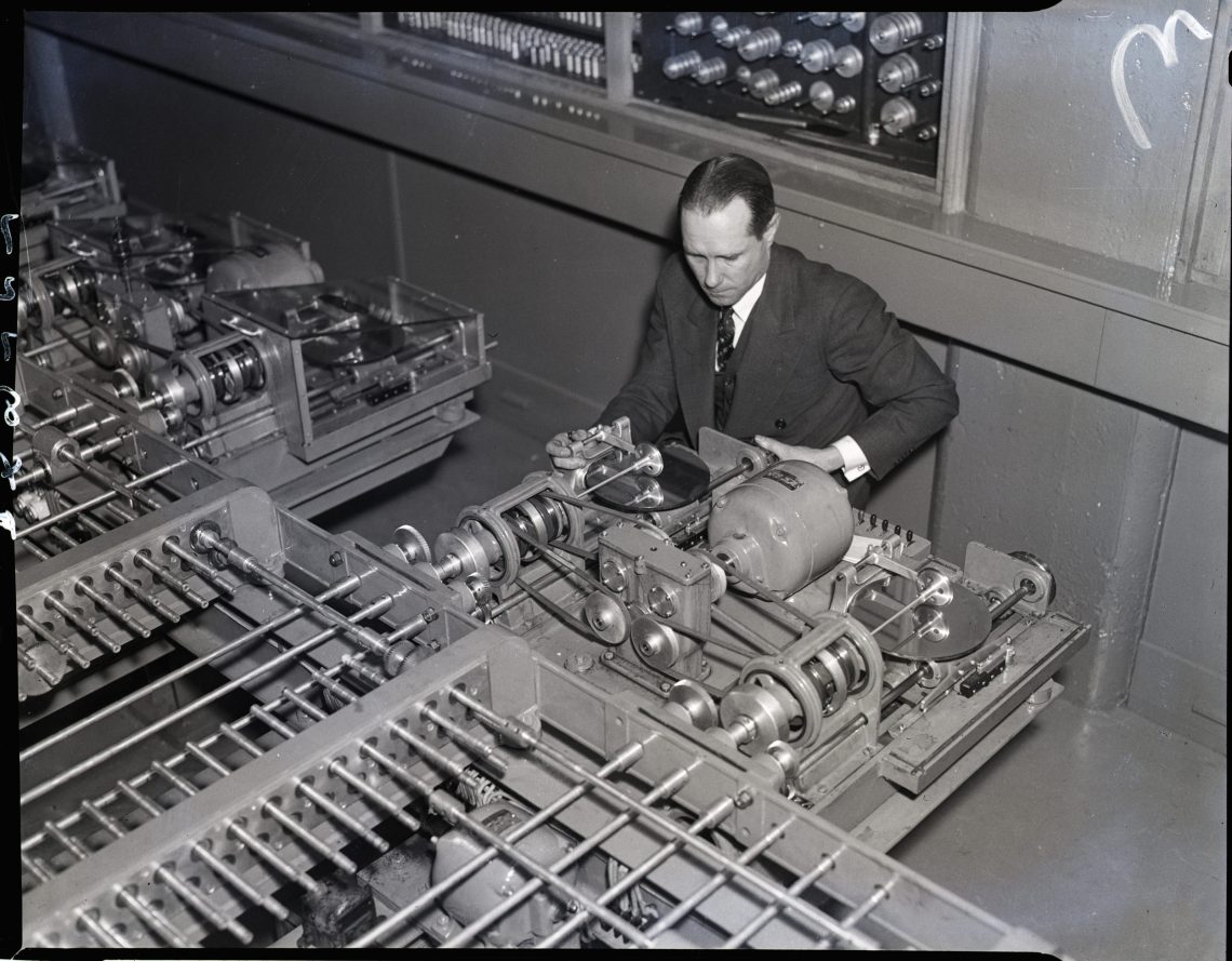 Ein Mann sitzt vor einer Maschine mit vielen Kompressoren. Schwarzweiß Foto, Draufsicht.