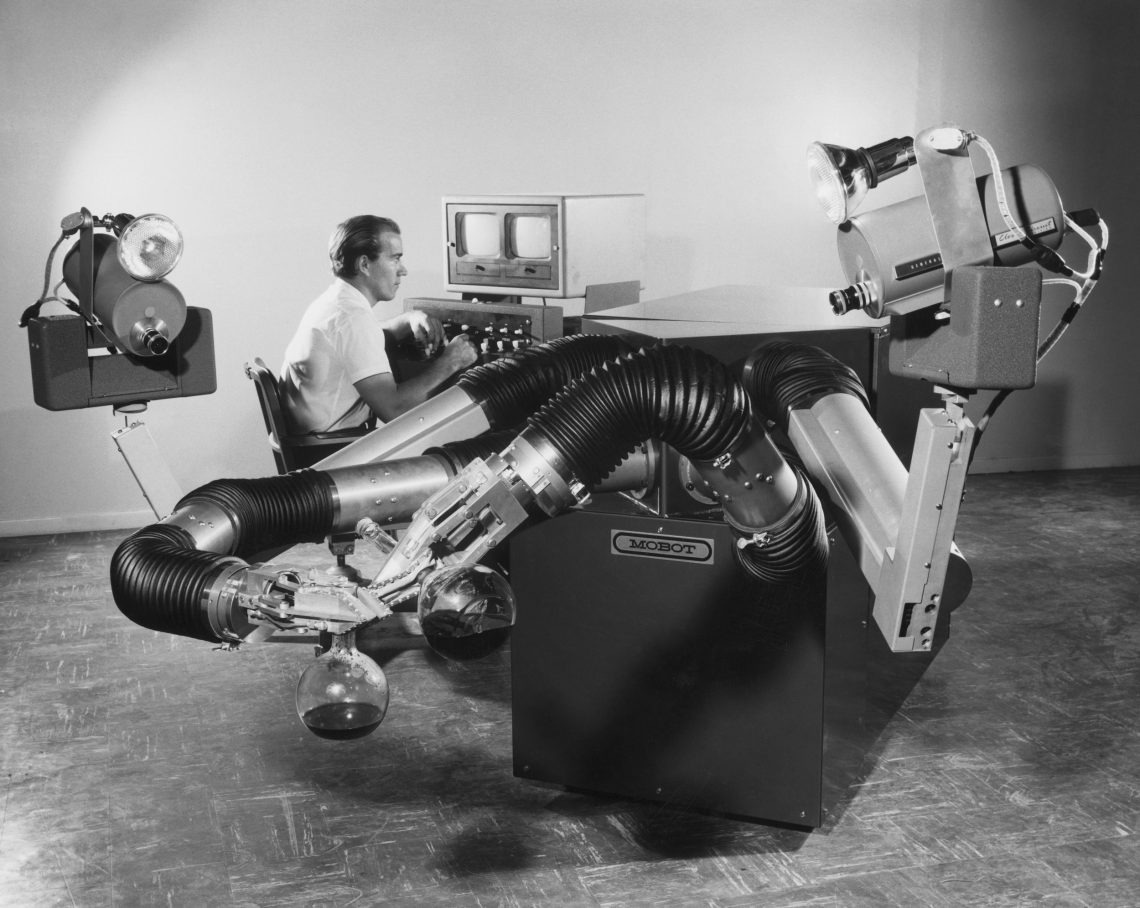 Schwarzweiß Foto eines Mannes, der in einem Bildschirm blickt. Er sitzt inmitten einer Maschine mit Armen wie Staubsaugerschläuche.
