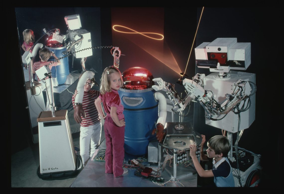 Foto einer Gruppe von Kindern, die zwei Roboter, der eine tonnenförmig, der andere eckig, unter die Lupe nimmt. Ein Mädchen in der Mitte stehend, dreht sich zur Kamera um.