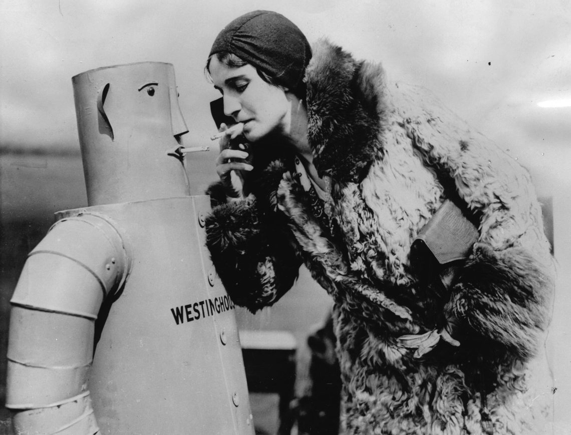 Eine Frau einem Pelzmantel mit Clutch unter dem einen Arm beugt sich zu einem Roboter, der ebenfalls eine Zigarette im Mund hat hinunter, wie um die Zigarette wieder zu entzünden.
