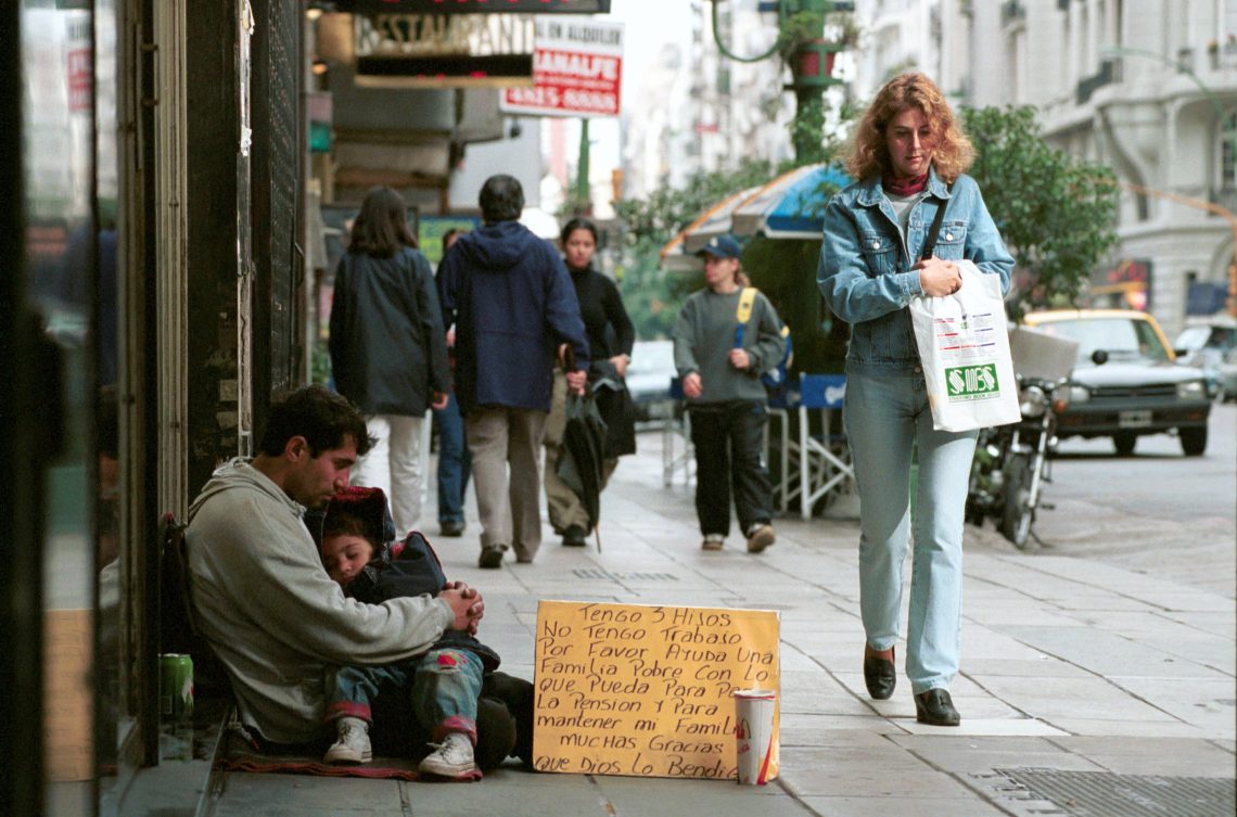 Ein Mann mit einem Kind im Arm sitzt auf dem Bürgrsteig einer Straße mit vielen Fußgängern und bettelt. Vor ihm steht ein Schild, auf dem steht, dass der Mann drei Kinder hat udn arbitslos ist.