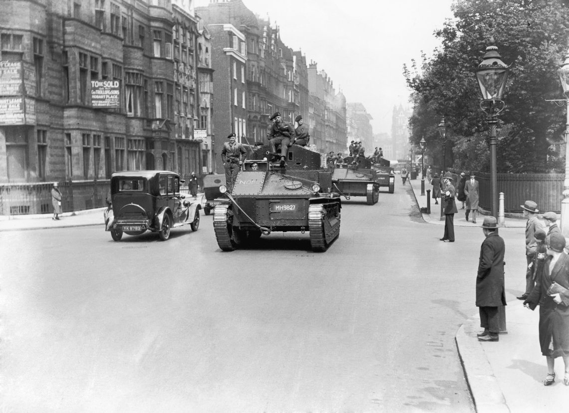 Foto von Panzern auf einer breiten Straße mit hohen Häusern und Bäumen auf einer Straßenseite. Auf dem Bürgersteig Menschen, die das Geschehen beobachten. Schwarzweiß Fotografie.