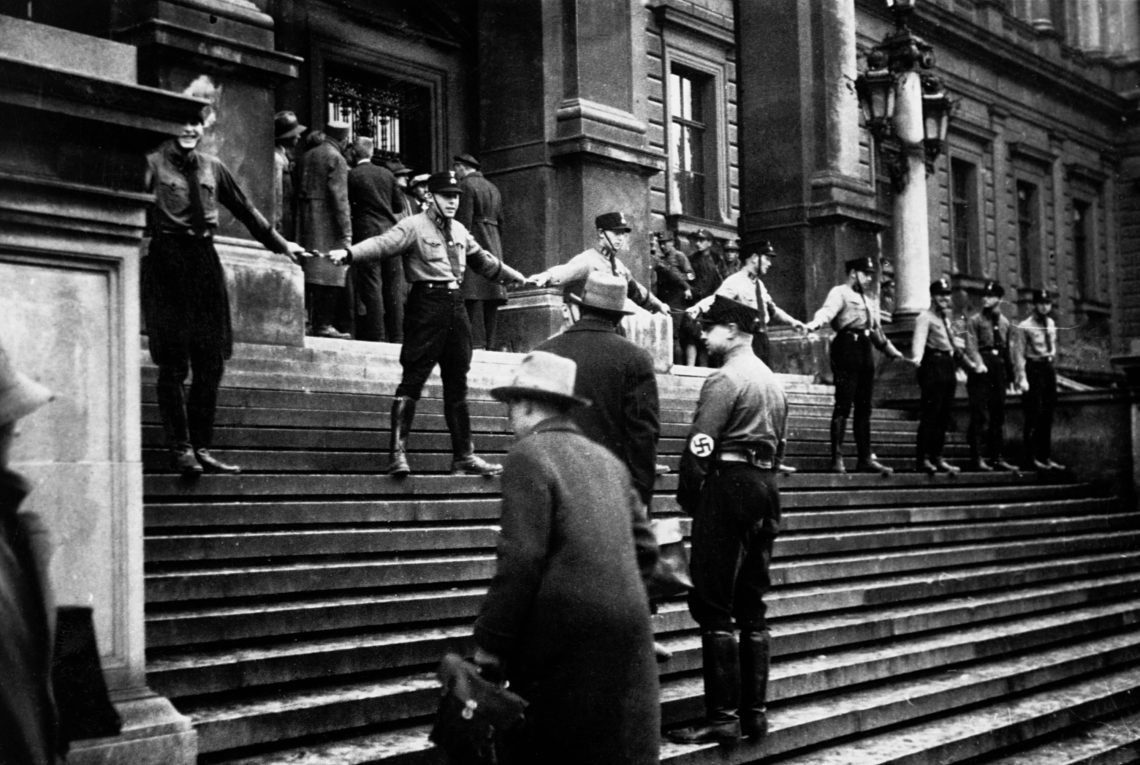 Auf den Stufen eines Gründerzeit-Gebäudes bilden Männer in Nazi-Uniformen eine Menschenkette und versperren den Eingang. Schwarzweiß Foto.