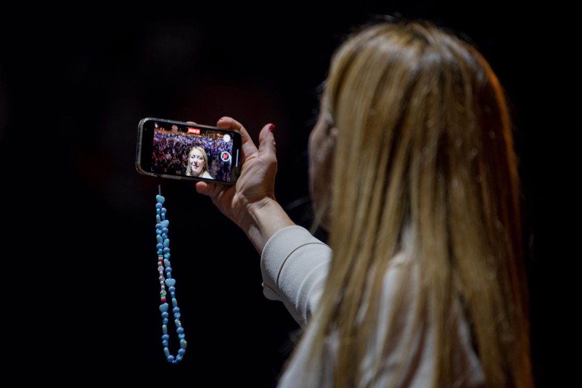 Populismus und Globalisierung: Rückenansicht von Giogia Meloni, die vor dunklem Hintergrund ein Selfie macht. In dem Selfie ist die Menge zu sehen zu der Meloni zuvor gesprochen hat.