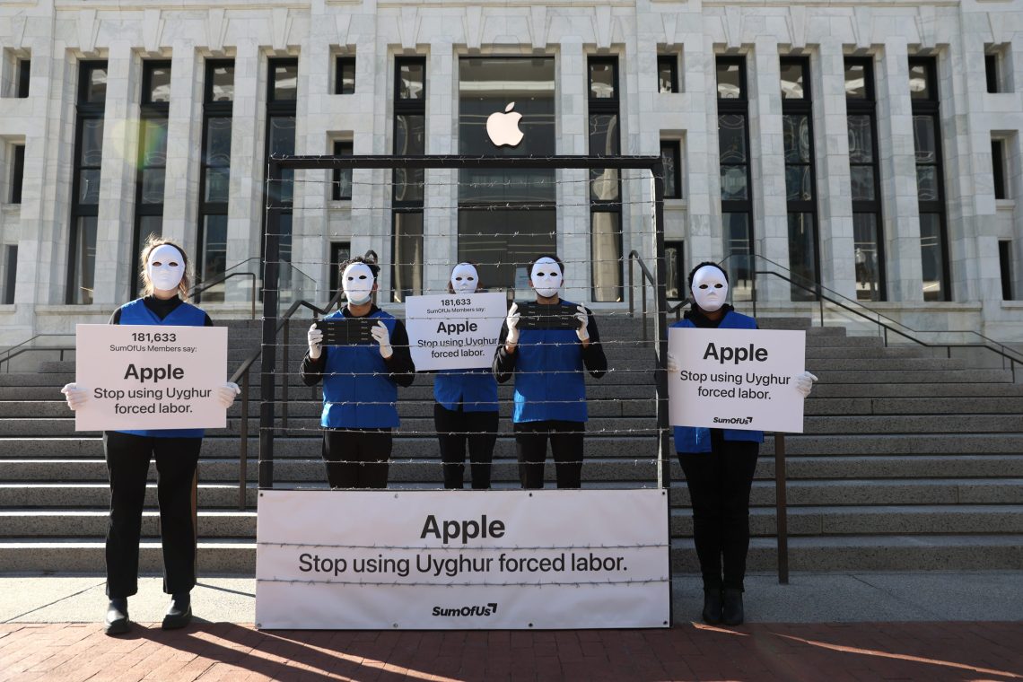Vor einem Gebäude mit dem Logo von Apple stehen Menschen mit weißen Michael-Myers-Gesichtsmasken und in blauen Westen mit Schildern auf denen steht „Apple, hör‘ auf, die Zwangsarbeit von Uiguren nutzen.“.