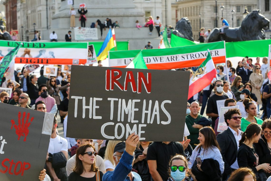 Der. Westen solidarisiert sich mit den Protesten im Iran: Foto einer Demonstration mit vielen Flaggen in den Farben des Iran. Ein Mann hält ein Plakat hoch mit der Aufschrift "Iran, deine Time ist abgelaufen".