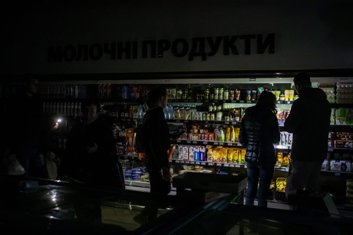 Foto eines Supermarktes, bei dem nur ein Kühlregal beleuchtet ist. Man kann schemenhaft die Kunden erkennen. Im Vordergund ist eine Kasse, über dem Regal steht etwas in kyrillischer Schrift.