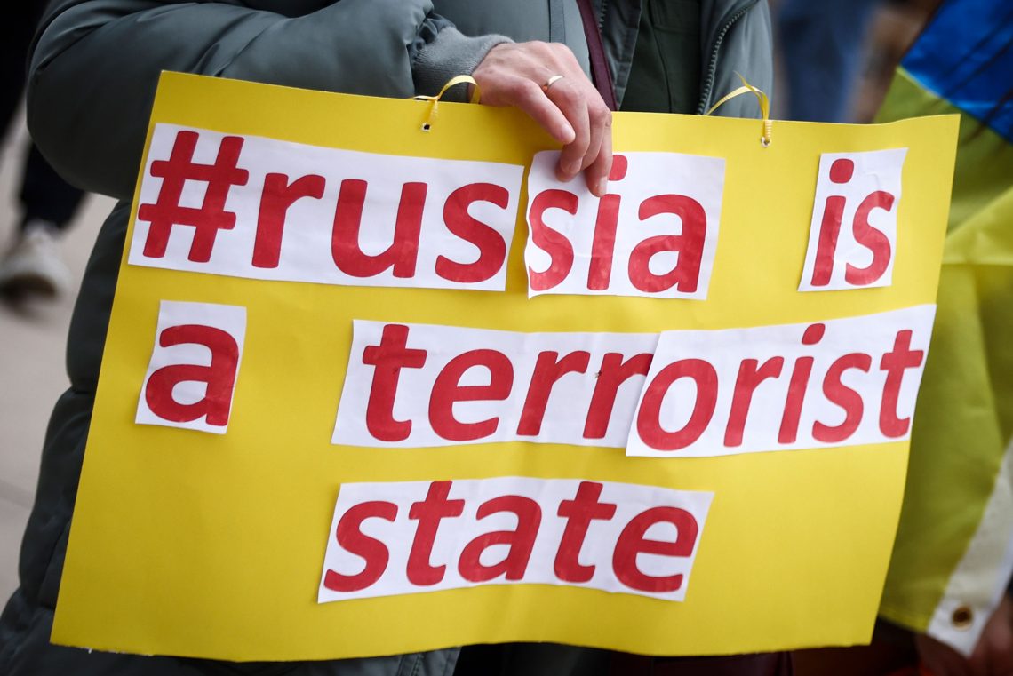 Foto eines gelben Plakates mit roter Aufschrift, das von einer Frau gehalten wir. Auf dem Plakat steht auf englisch Russland ist ein Terrorstaat. Das Foto soll einen Beitrag über die Legitimität von Tyrannenmord illustrieren.