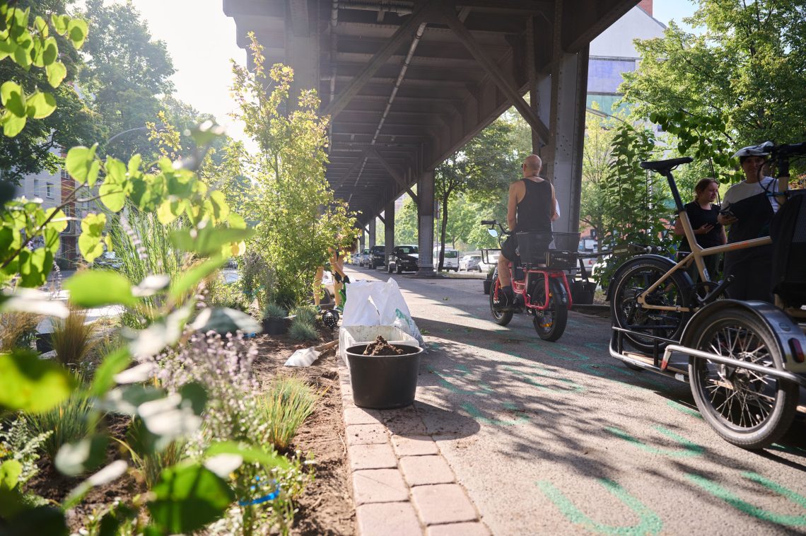 Ein Fahrradweg mit Beeten unter einer U-Bahntrasse im Sonnenschein. Illustration zu einem Beitrag über die Naivität der Energiewende.