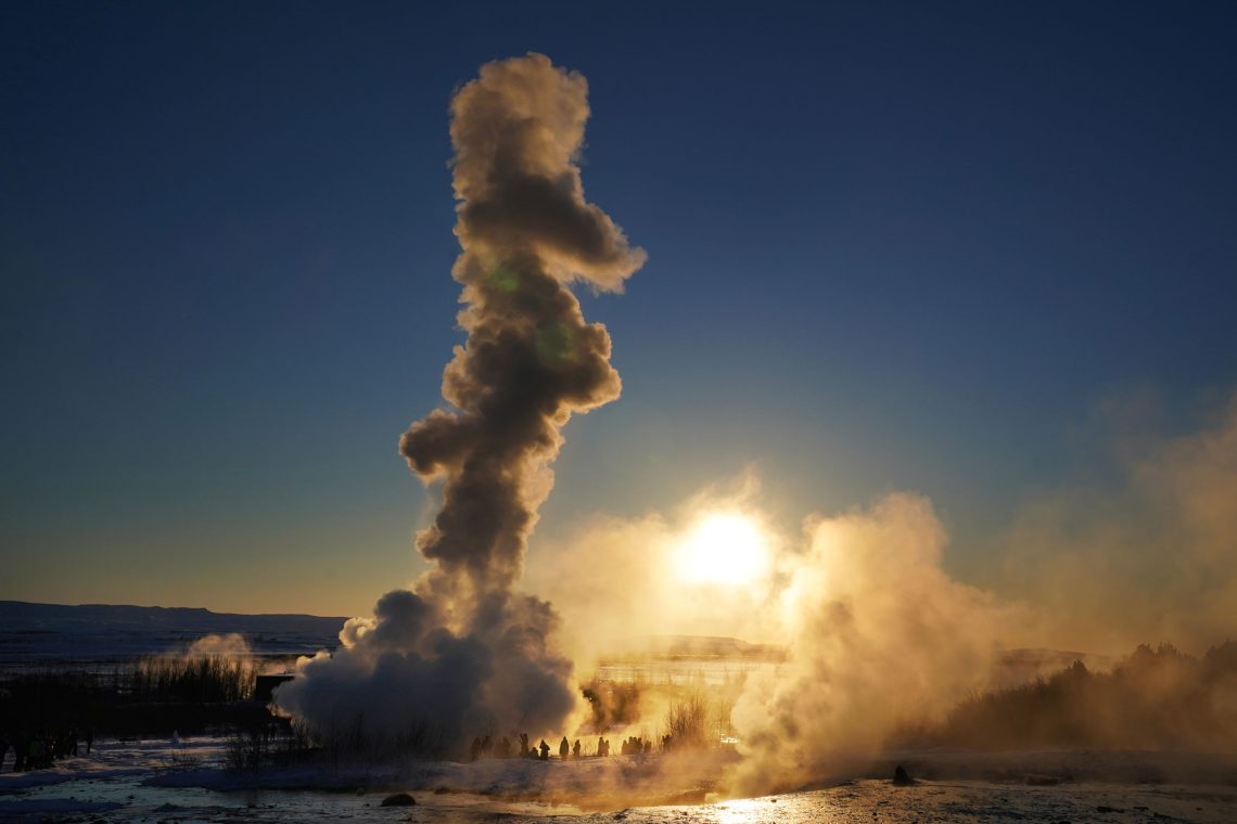 Foto eines Geysirs bei Sonnenaufgang in einer Schneelandschaft. Der Wasserdampf steigt so hoch, dass die Menschen, die sich das Schauspiel anschauen ganz klein im Vergleich sind sind. Das Bild soll die Wärme und Energie illustrieren, die durch Geothermie vorhanden ist.