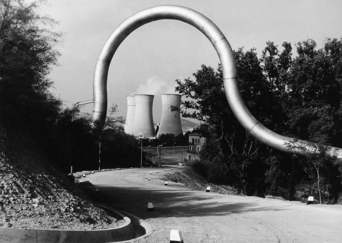 Schwarzweiß Foto eines großen gebogenen Rohres über einer Straße. Durch die Schlaufe sieht man im Hintergrund drei große Kühltürme, aus denen Wasserdampf aufsteigt. In dem Bild geht es um Geothermie.