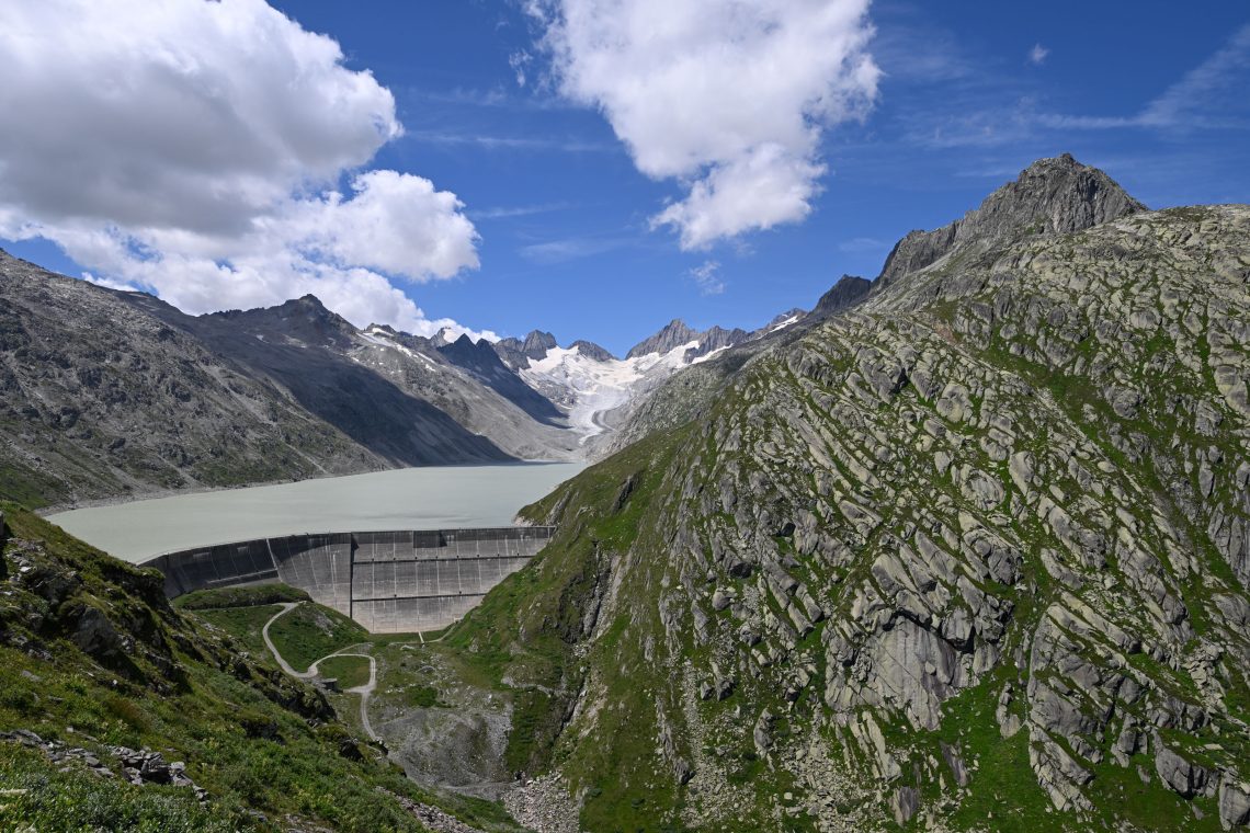 Ansicht einer Berglandschaft in der Schweiz mit einem Gletscher im Hintergrund und einem Stausee mit großer Staumauer im Vordergrund.