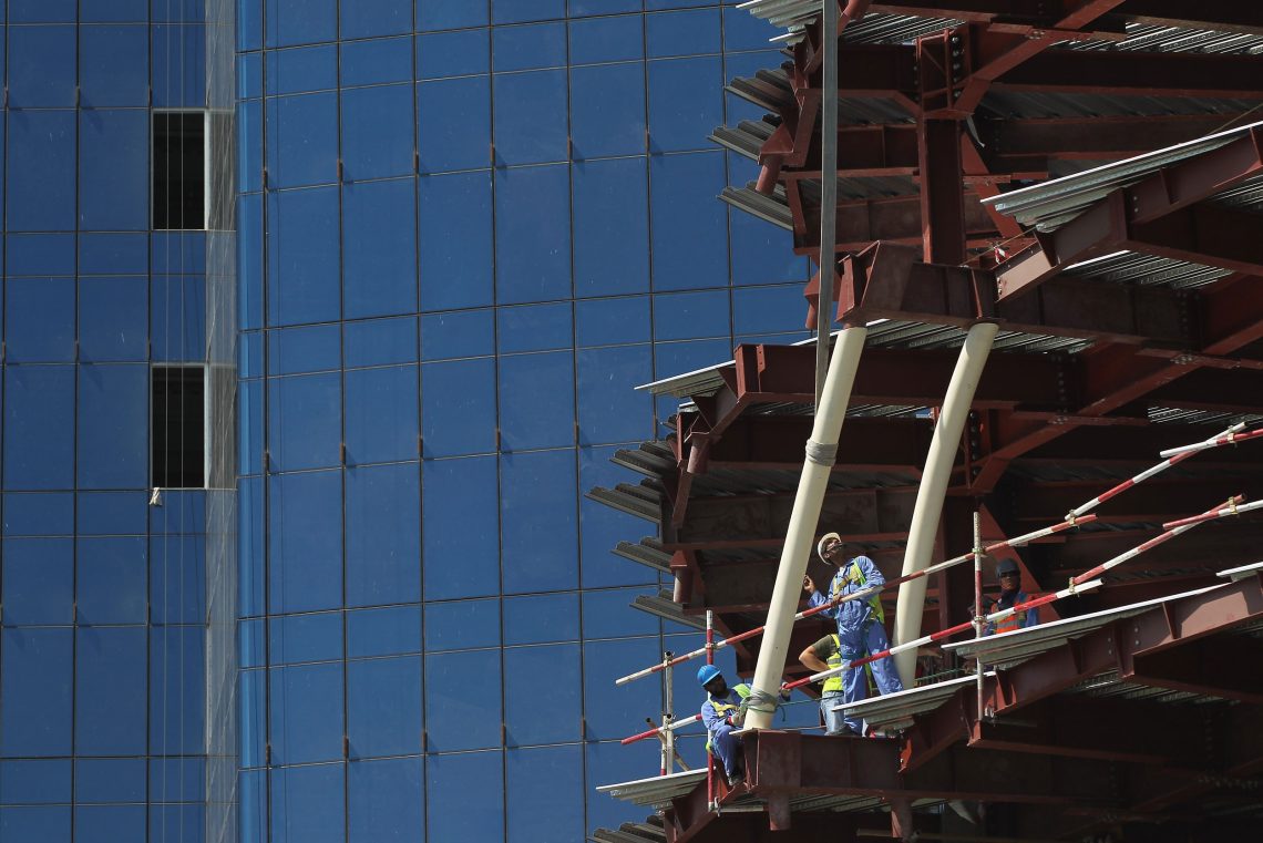 Foto eines hohen Gebäudes ohne Außenmauern mit Arbeitern. Im Hintergrund eine große Glasfassade. Das Bild soll das Verhältnis von Katar und dem Westen illustrieren.