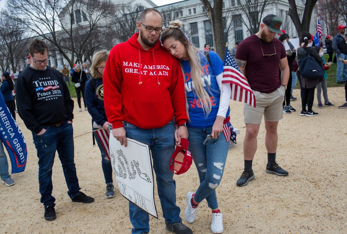 Foto einer Gruppe von Menschen die mit geschlossenen Augen und gesenkten Köpfen stehen. Sie halten Schilder in der Hand auf denen steht "Stop the Steal" und Trump sowie T-Shirts mit der Aufschrift "Make America great again".