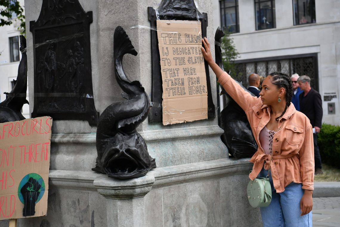 Eine schwarze junge Frau steht neben dem Sockel einer Statue und hält einen Karton mit der Aufschrift "This Place is dedicated to the Slaves that were taken from their homes". Das Bild soll einen Beotrag über Cancel Culture Panik illustrieren.