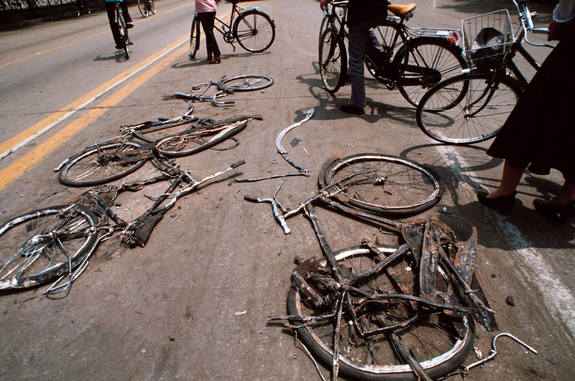 Auf einer Asphalt-Straße sind Fahrräder zu sehen, die platt gefahren wurden. Menschen mit Fahrrädern stehen und sehen sich das an. Das Foto wurde nach den Protesten 1989 in China aufgenommen. Die Proteste waren damit beendet.