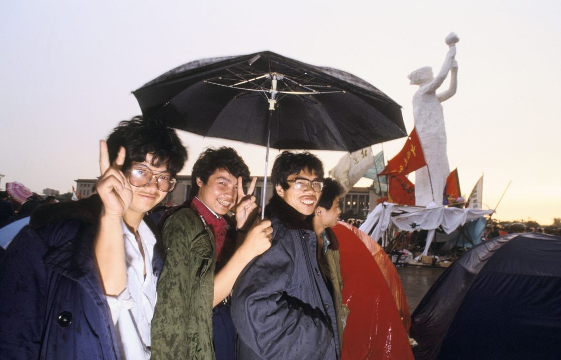 Foto von vier Jugendichen, einer mit Regenschrim, die in die Kamera lachen, wobei einer von ihnen ein Victory-Zeichen macht. Das Foto wurde bei Protesten 1989 in China aufgenmmen.
