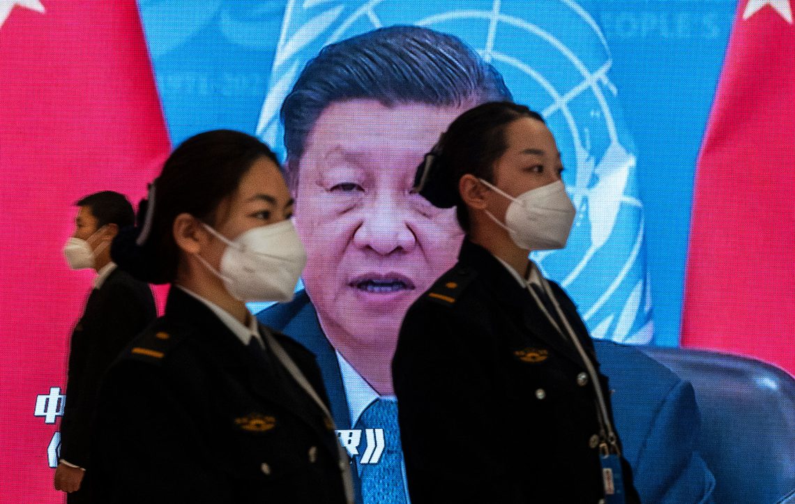 Foto von Xi Jinping auf einer Videoleinwand, im Vordergrund stehen zwei uniformierte Frauen mit Gesichtsmasken. Das Foto begleitet einen Beitrag über China und die chinesische Wirtschaft.