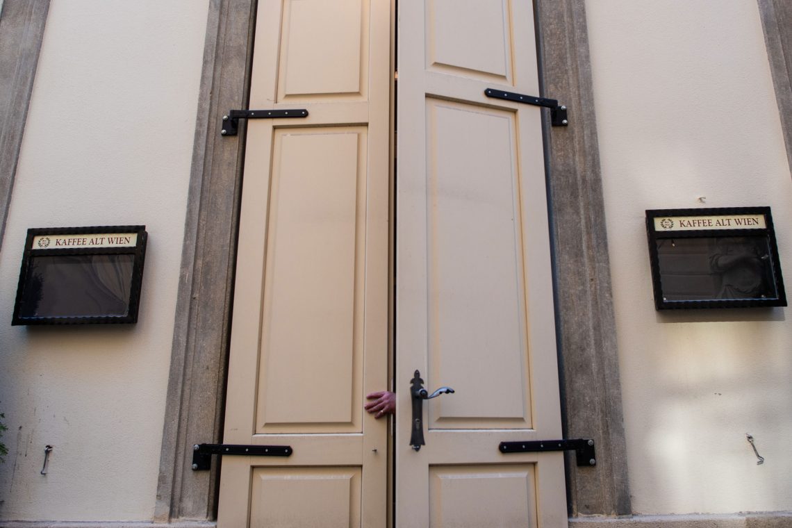 Eine hohe Tür mit Flügeln wird von innen zugezogen. Neben der Tür hängen leere Auslagen für Speisekarten mit der Aufschrift Kaffee Alt Wien. Das Bild illustriert einen Beitrag über die Folgen der Corona-Maßnahmen in Österreich.