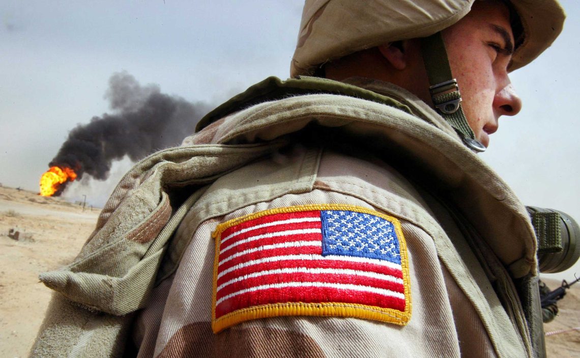 Nahaufnahme eines amerikanischen Soldaten mit US-Flagge an der Schulter während des Irakkriegs 2003. Im Hintergrund lodert ein Feuer und dichter, schwarzer rauch steigt über einem brennenden Ölfeld auf.