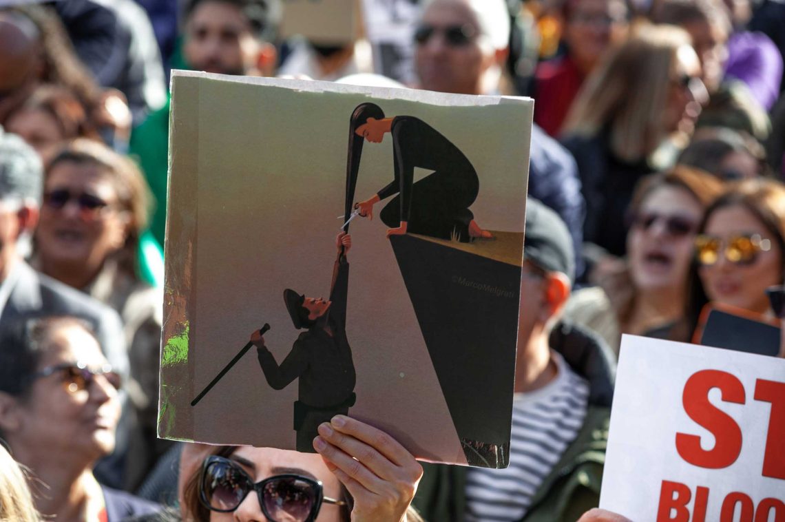 Den Haag: Eine Demonstrantin für Frauenrechte im Iran hält ein Schild hoch. Darauf ist eine Zeichnung zu sehen, von einem Vertreter der Moralpolizei mit Schlagstock, der über einem Abgrund hängt und sich an den Haaren einer knienden Frau festhält, die im Begriff ist ihre Haare abzuschneiden.