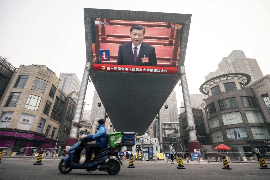 Ein Zusteller fährt auf einem Motorrad an einem Bildschirm vorbei, auf dem eine Live-Nachrichtenübertragung des chinesischen Präsidenten Xi Jinping auf dem Nationalen Volkskongress in Peking zu sehen ist.
