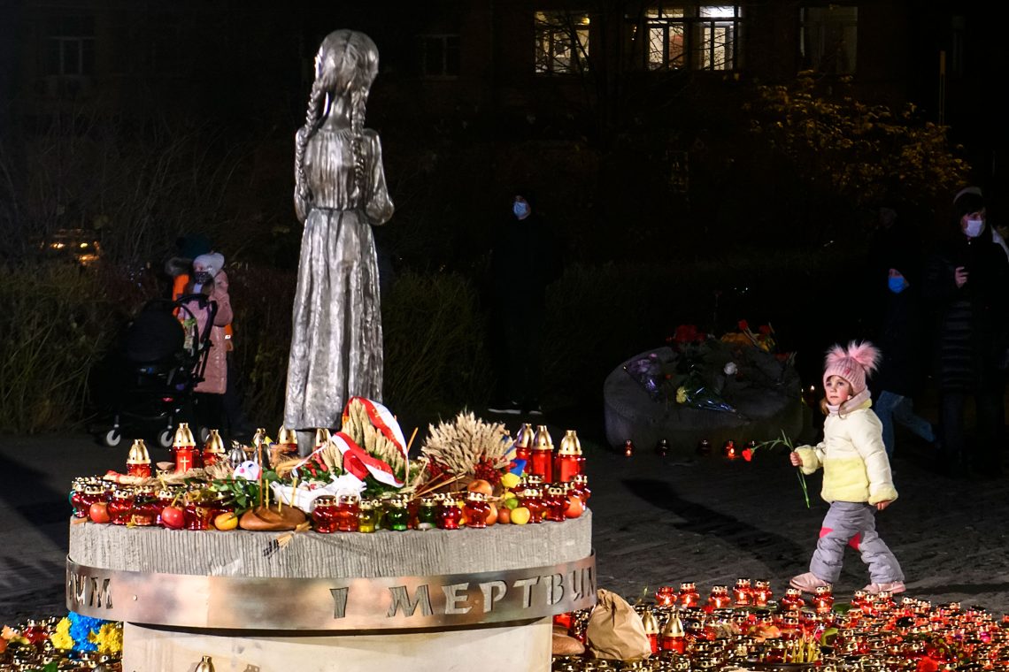 Foto der Bronzeskulptur eine Mädchens mit langen Zöpfen zu deren Füßen auf dem Sockel viele Kerzen und Weizenbüschel, Äpfel, Fahnen und Blumen liegen. Das Bild zeigt das Gedenken an den Holodomor, der international nicht als Völkermord gilt.