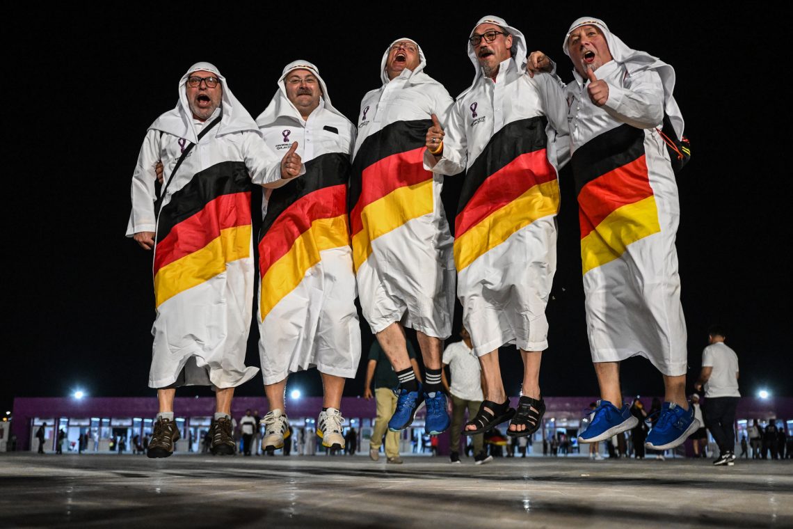 Foto von fünf Männern mittleren Alters, die mit einem weißen Gewand, ähnlich wie ein Thawb und eienr Kopfbedeckung, die einem Igal ähnelt, hochspringen. Die wadenlangen Gewänder sind mti einer Schärpe in den Nationalfarben von Deutschlands bedruckt.