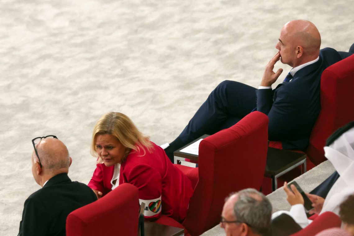 Foto einer Sitzreihe mit großen roten Sesseln. Die Innenministerin von Deutschland trägt einen roten Anzug mit einer Armbinde einem Herz in Regenbogenfarben. Sie beugt sich zu einem Mann mit Glatze, Bernd Neuendorf, DFB-Präsident. Neben der Frau sitzt rechts ein weiterer Mann mit Glatze, der, eine Hand am Mund, nachdenklich nach vorne blickt.