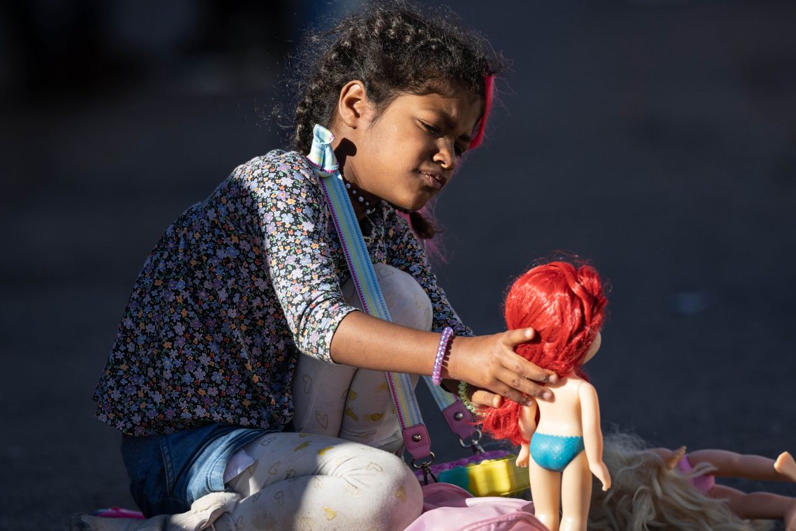 Ein Mädchen mit ernstem Gesichtsausdruck sitzt im Sonnenlicht und streicht über das rote Haar einer Puppe. Das Bild illustriert einen Beitrag über Migration, Bevölkerung und Überalterung.