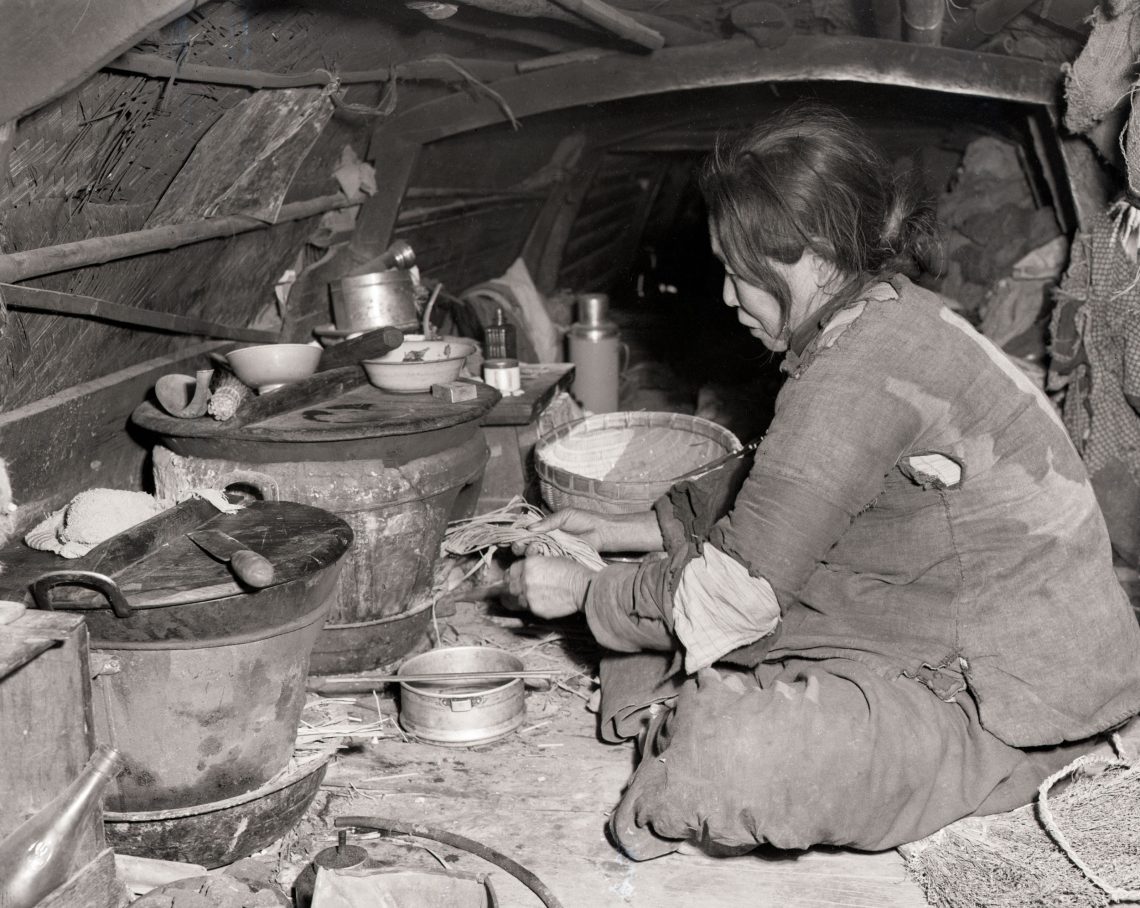 Eine Frau sitzt im Schneidersitz auf dem Erdboden in einer provisorischen niedrigen Behausung und heizt einen Ofen mit Stroh. In dem Beitrag zu dem Bild geht es um Politik und China.